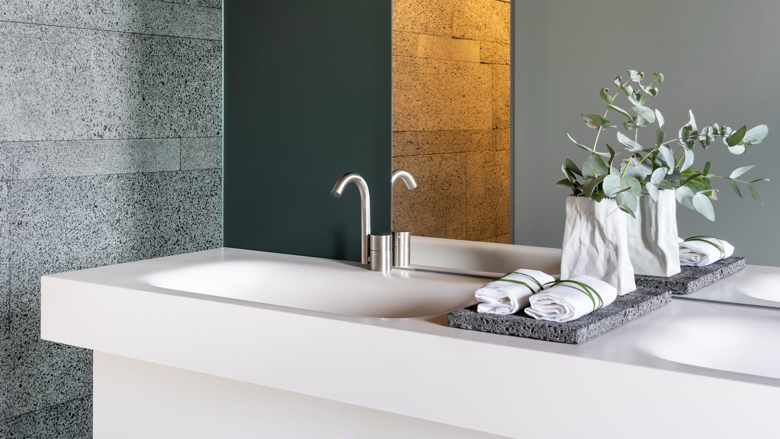 Encimera de baño del Hotel Zash con griferia Fantini en acero, pared de piedra volcánica gris y panel dorado al fondo.
