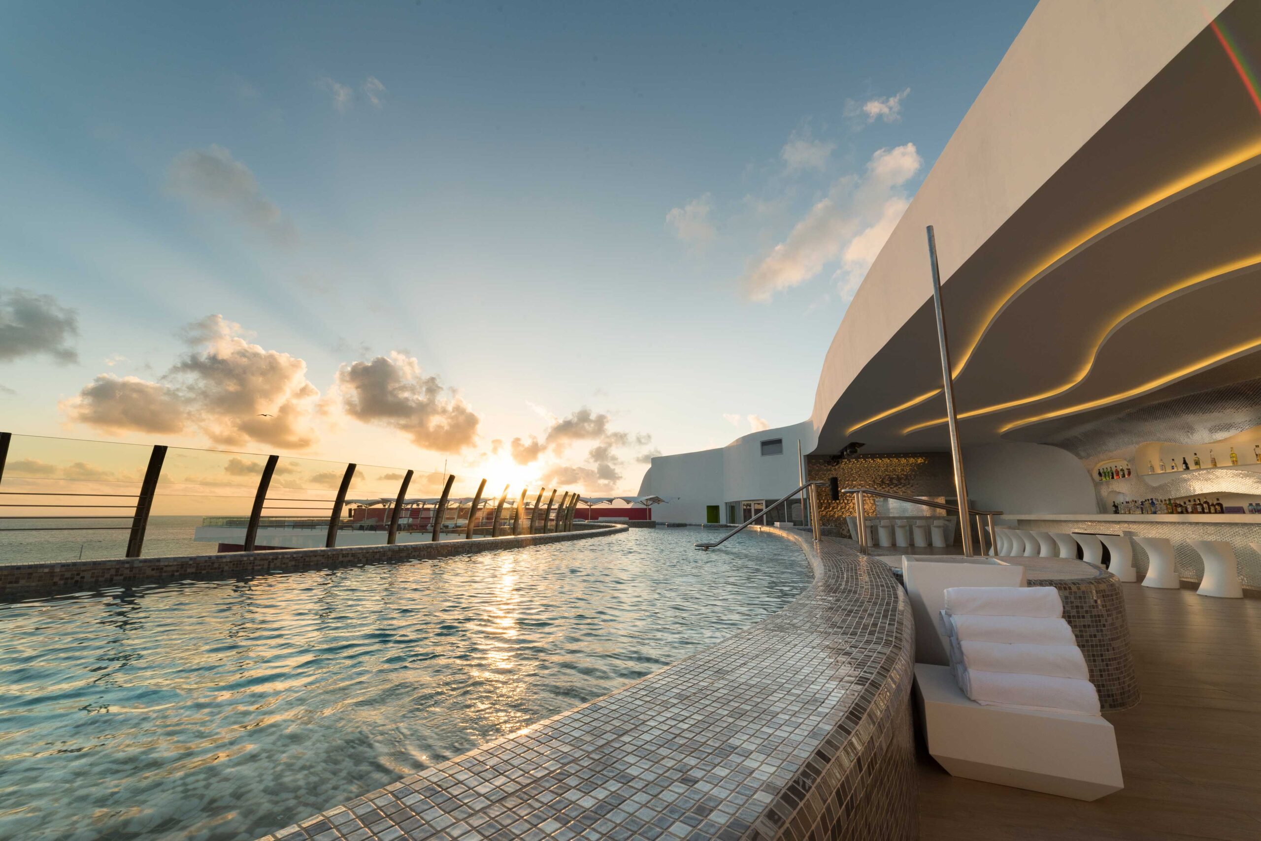 Piscina infinita de un skybar al atardecer, con el borde y el vaso revestidos de mosaico vítreo y el mar al fondo.